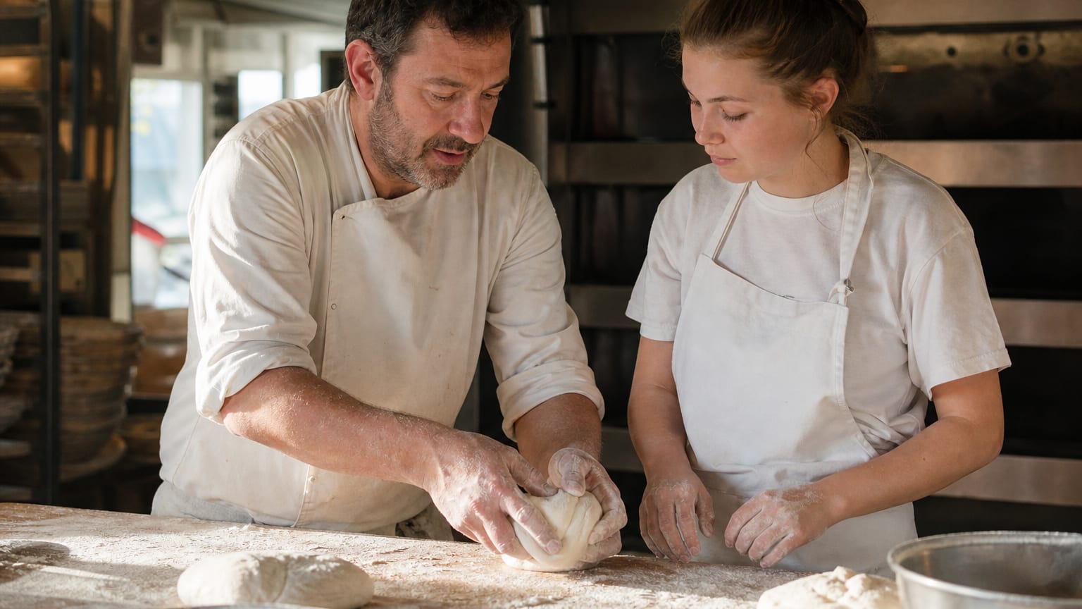 Un artisan boulanger montre un geste à son apprentie dans le fournil d’une boulangerie de quartier